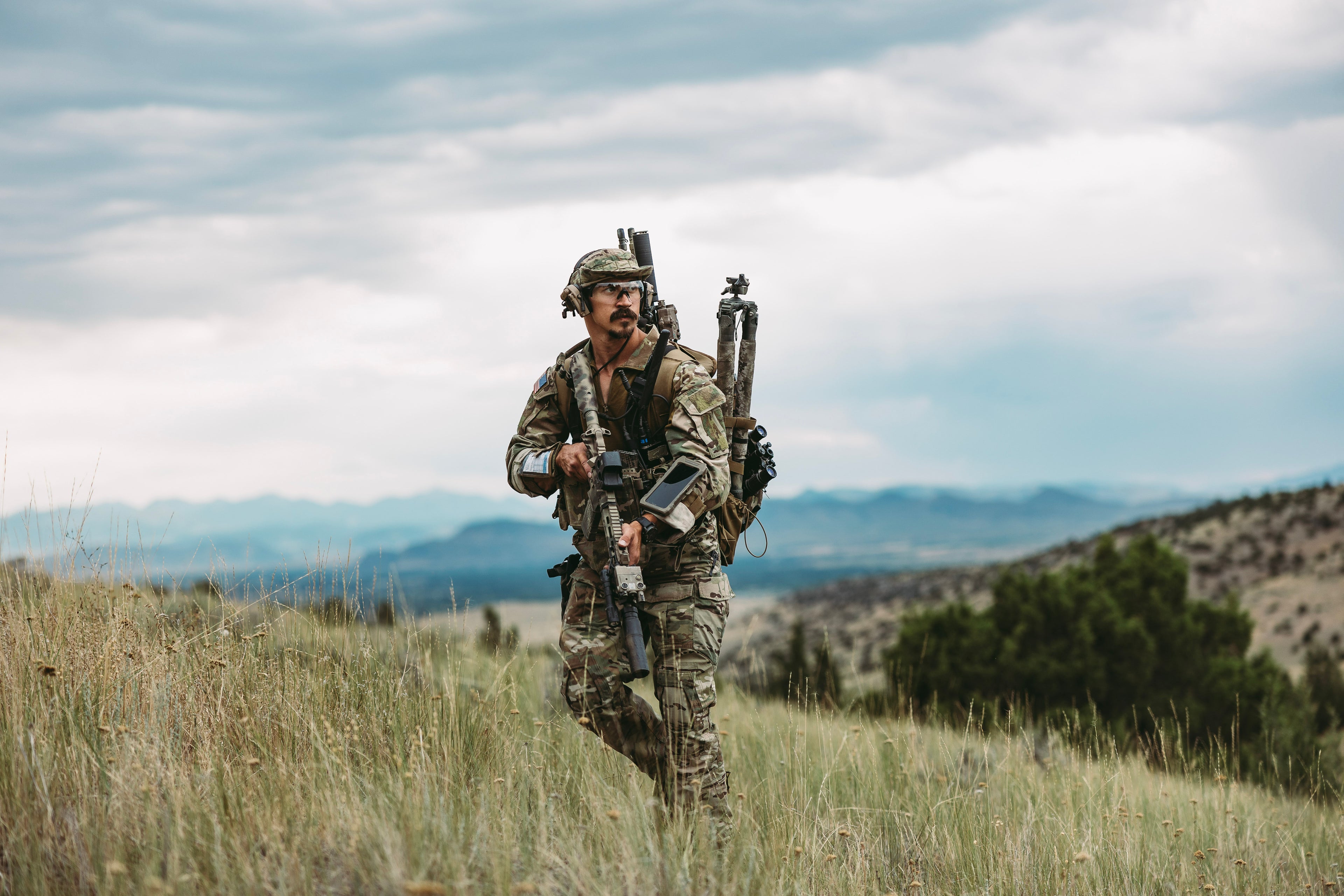 Person in camouflage walking through a grassy field with mountains in the background