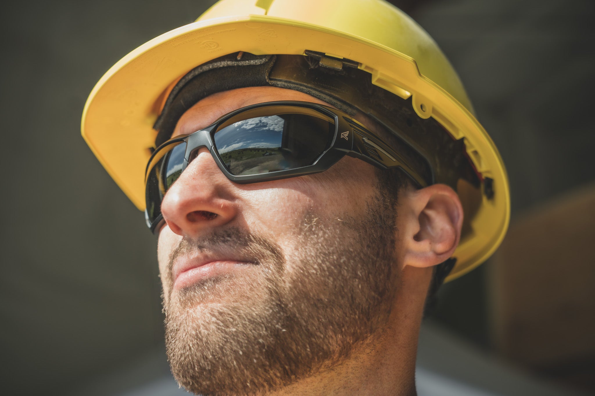 man wearing a yellow hard hat and tinted black safety glasses with blurred background