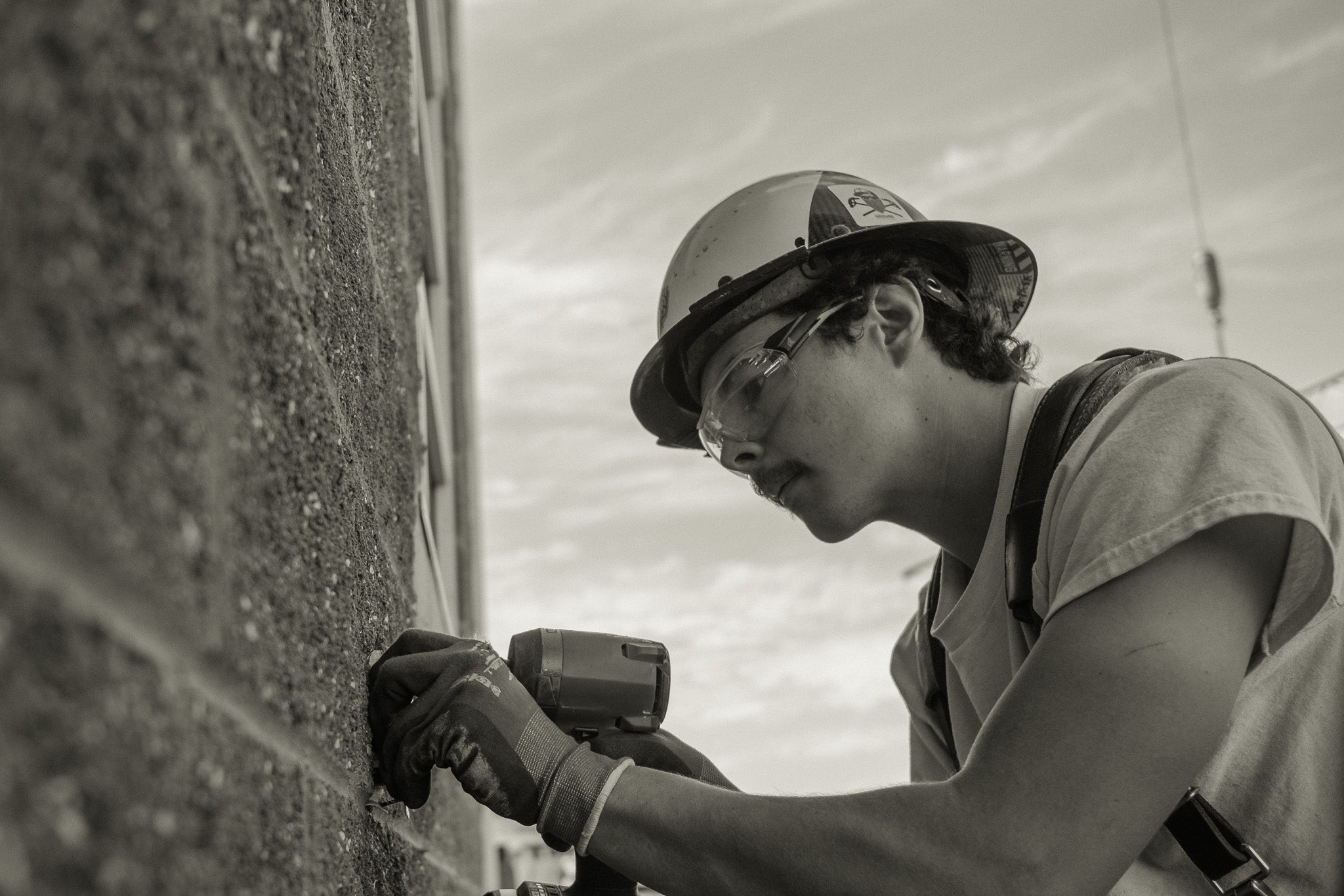 Person wearing a hard hat and safety glasses using a power tool on a wall.