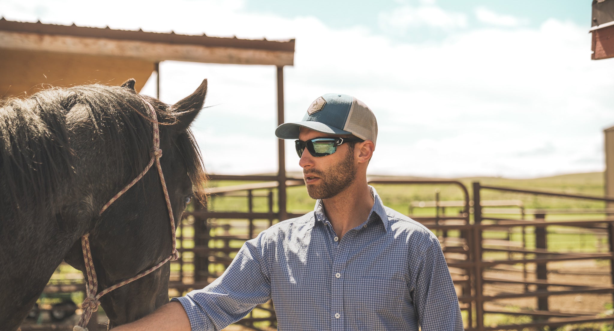 Man wearing safety sunglasses and a cap standing next to a horse in an outdoor setting