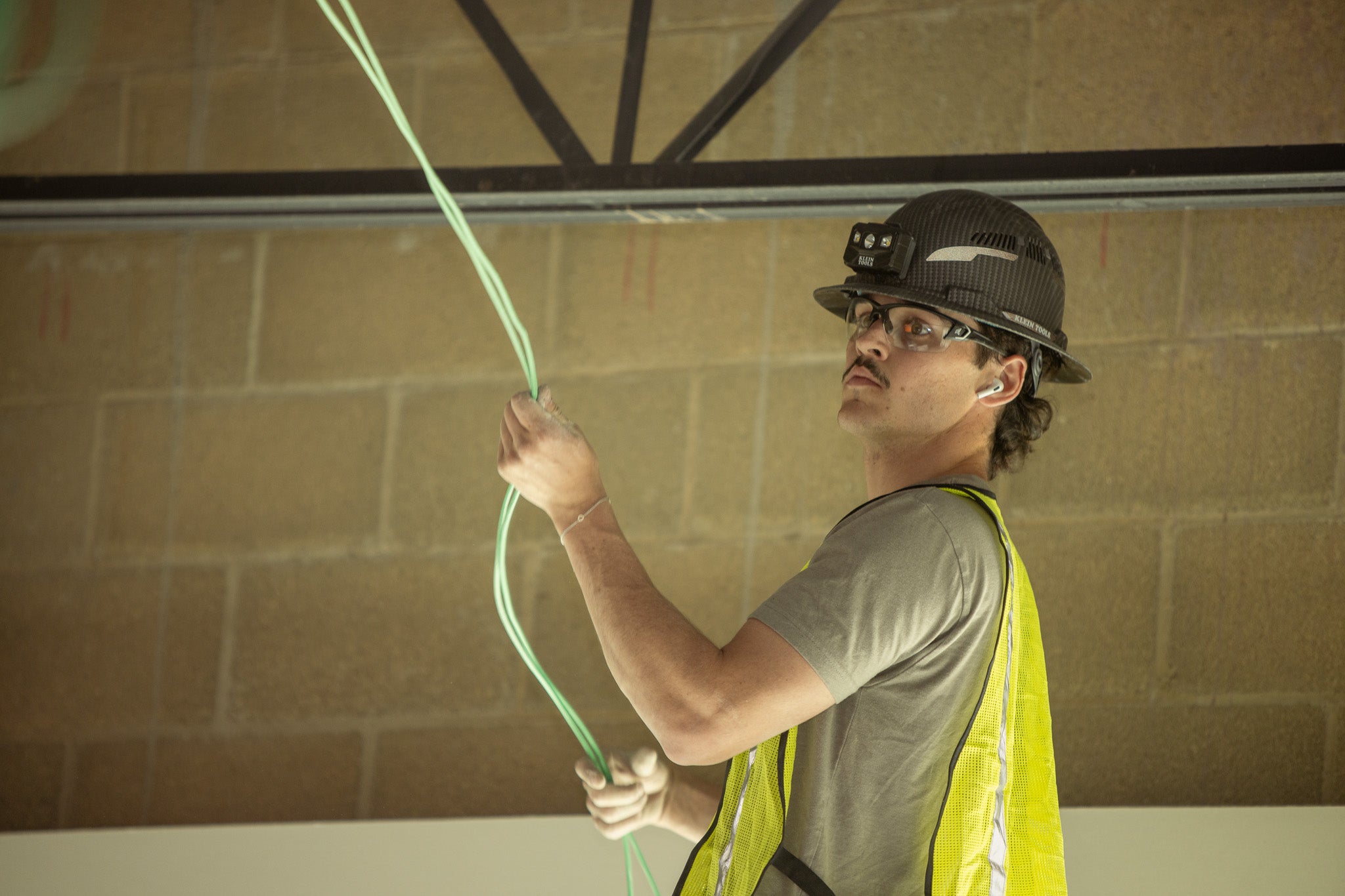 Person in a yellow safety vest, glasses and hard hat holding green cable against a brick wall.