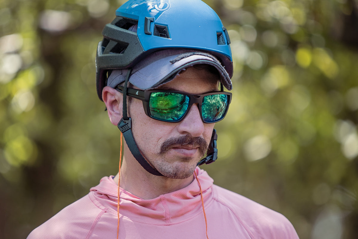 A close-up of a man wearing a blue mountain bike helmet, mirrored green sunglasses, and a pink long-sleeve athletic jersey, set against a blurred green outdoor background.