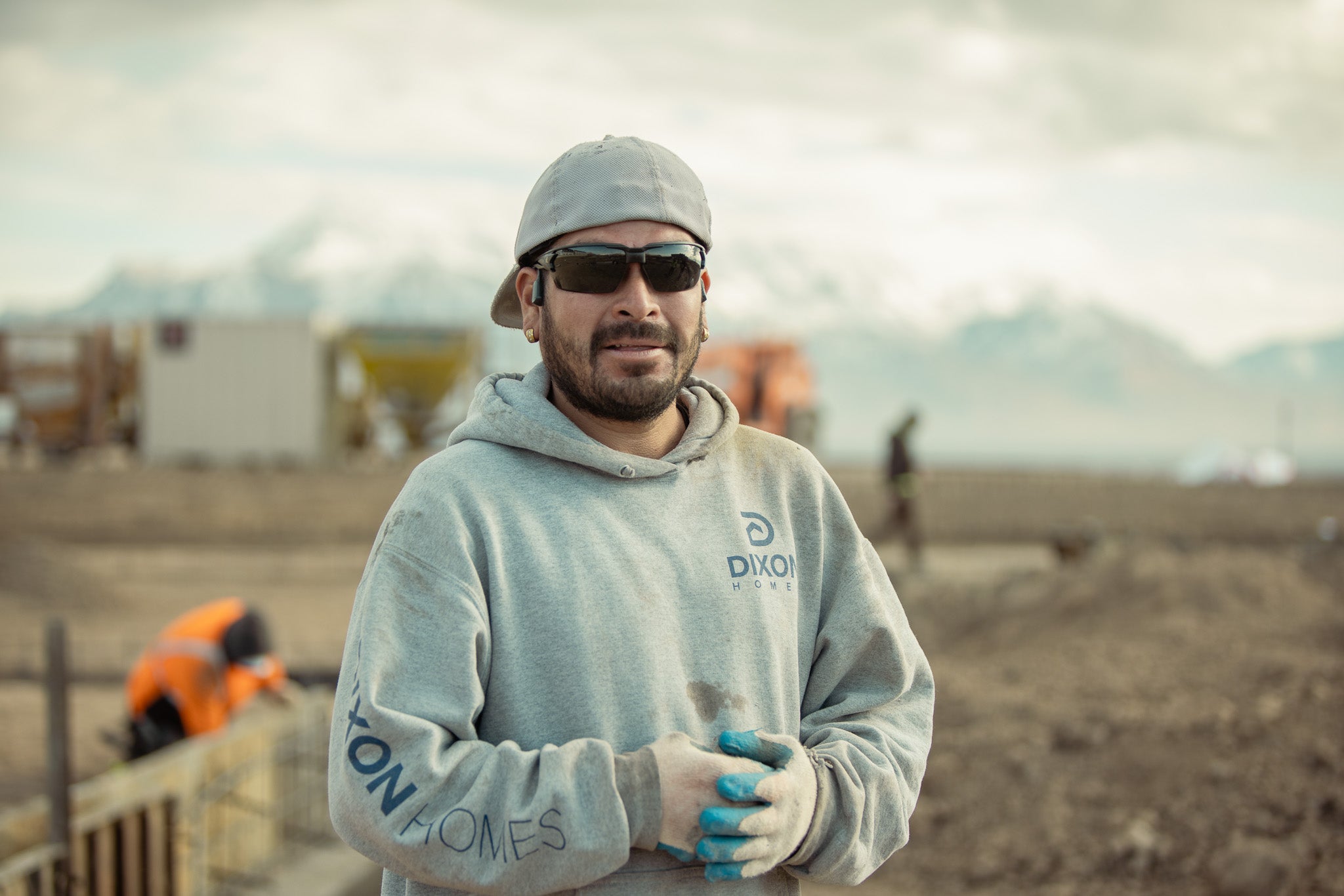 Man wearing a gray hoodie with a logo, safety glasses, and gloves standing on a construction site.