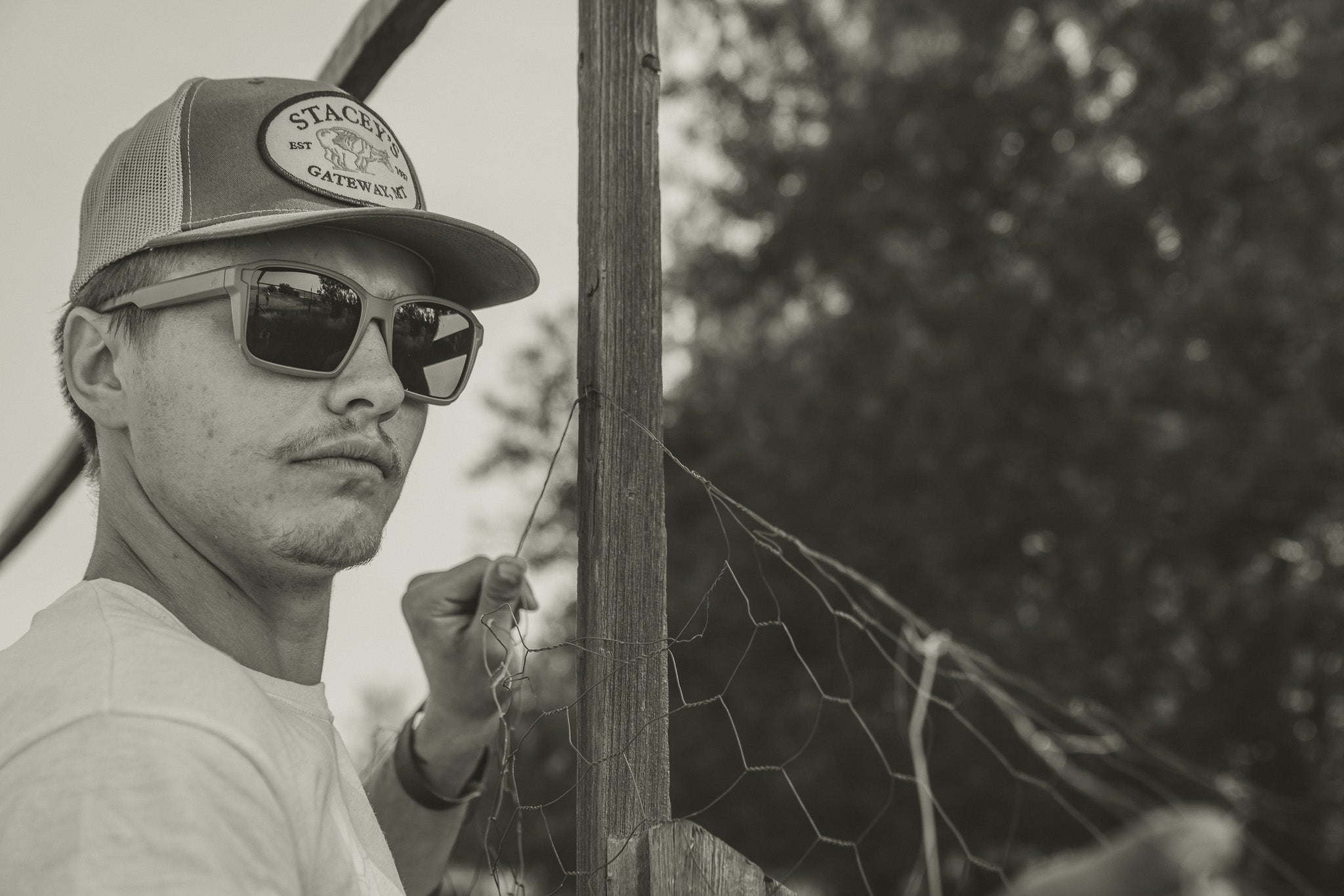 Person wearing sunglasses and a cap, standing next to a fence with trees in the background