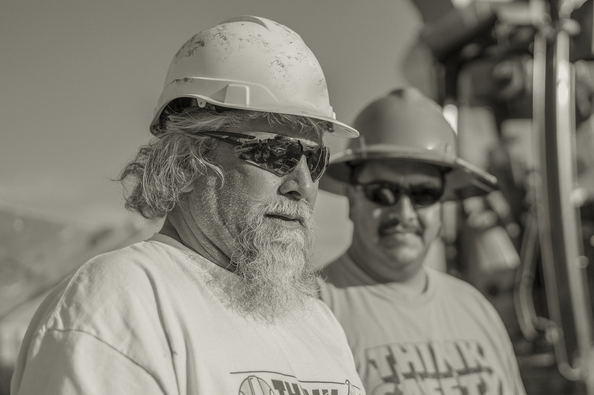 Two men wearing hard hats and sunglasses in a construction or industrial setting.