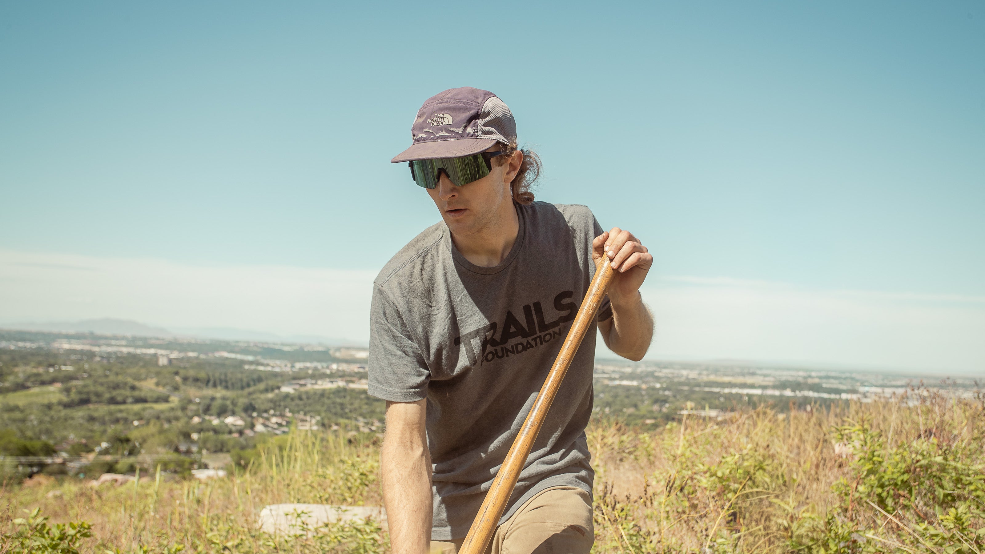 A man wearing a gray t-shirt with 'RAILS FOUNDATION' printed on the front, a purple cap, and sunglasses, holding a wooden pole outdoors on a grassy hill with a scenic valley in the background.