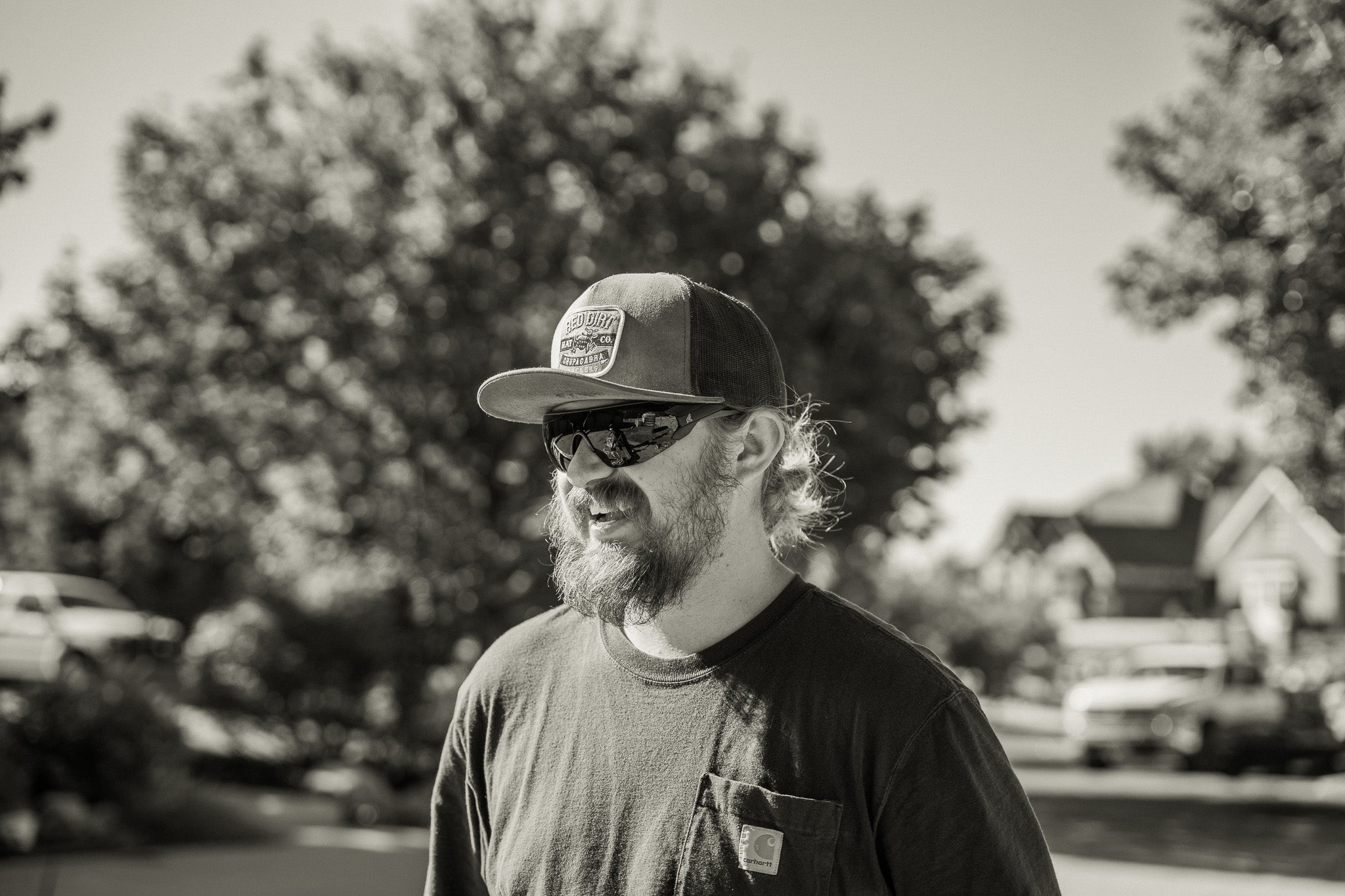 Man wearing a cap and sunglasses outdoors with trees and buildings in the background