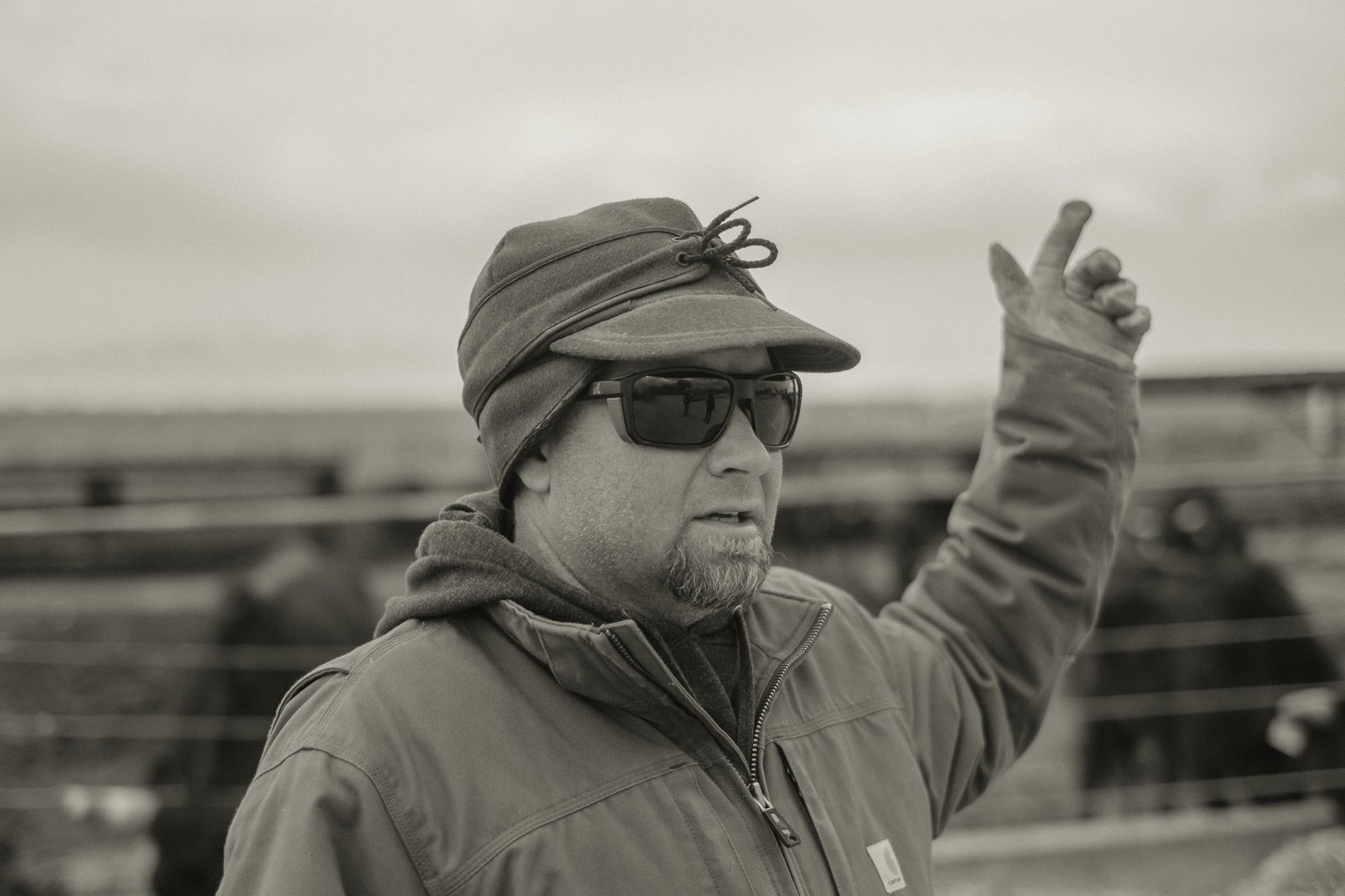 Man wearing a cap, sunglasses, and jacket pointing with a cloudy sky background
