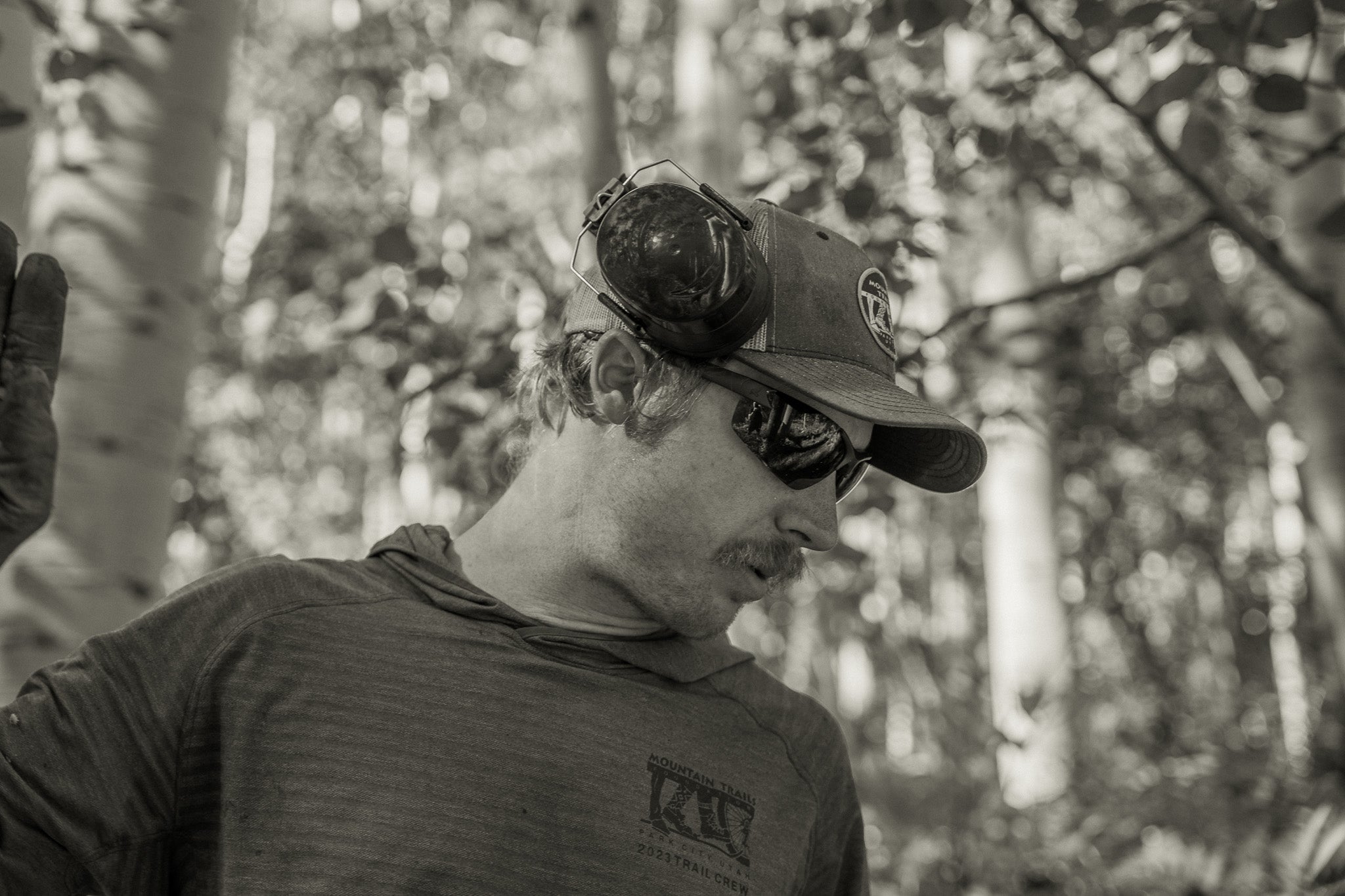 man wearing a baseball cap and black safety sunglasses holding up his hand in an outdoor setting. 